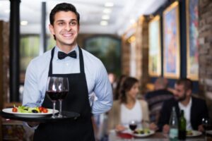 coppia seduta al tavolo di un ristorante e cameriere che sorride con in mano un vassoio